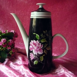 Könnte beinhalten: Eine hohe, schwarze Keramik-Kaffeekanne mit weißem Ausguss und Griff. Die Kanne ist mit einem rosa-weißen Blumendesign verziert. Der Deckel hat einen weißen Deckel und schwarz-weiße Streifen. Im Hintergrund befindet sich ein Strauß rosa Blüten.