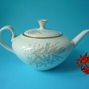 Op de afbeelding: Een witte porseleinen theepot met een taps toelopend lichaam, een gebogen handvat en een bijpassend deksel met een goudkleurige rand. Het oppervlak is versierd met een gestileerd botanisch patroon in blauw, geel en grijstinten en een tuit.