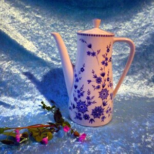 Op de afbeelding: Een hoge, witte keramische koffiepot met een deksel en handvat, versierd met blauwe bloempatronen. De pot heeft een lange tuit en staat tegen een blauwe achtergrond. Een takje bloemen met roze bloesems ligt in de buurt.