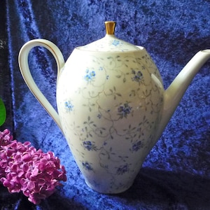 Puede incluir: Una tetera de porcelana blanca con tapa, decorada con un delicado estampado floral azul. El asa y el pico son curvos, y la tapa tiene un pomo dorado. Una ramita de flores moradas y hojas verdes en primer plano.