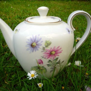 Peut inclure: Théière en porcelaine blanche ornée de fleurs violettes et roses. La théière a une bordure dorée et une anse incurvée. Elle est posée dans un lit d'herbe verte avec de petites fleurs blanches.