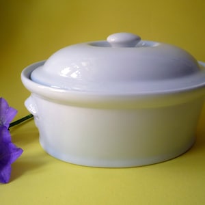 Op de afbeelding: Een witte keramische ovenschaal met een deksel en kleine handvatten. De schaal staat op een gele achtergrond, met paarse bloemen op de voorgrond.