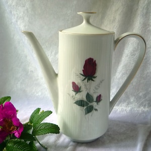 Op de afbeelding: Een hoge witte porseleinen koffiepot met een fijne ribbelstructuur en een centraal bloemmotief van een donkerrode roos met knoppen en groene bladeren. De pot is voorzien van een bijpassend deksel en heeft gouden randen.