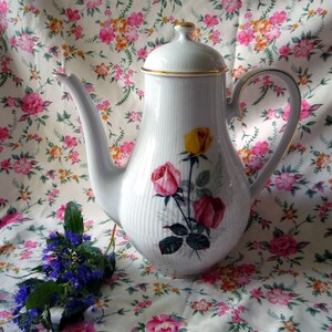 Könnte beinhalten: Eine weiße Kaffeekanne aus Porzellan mit gerippter Textur, passendem Deckel und goldfarbenem Rand. Der Körper ist mit einem Blumenmotiv aus einer gelben Rose und zwei rosa Rosen mit grünen Blättern verziert. Mit geschwungenem Henkel und Ausguss.