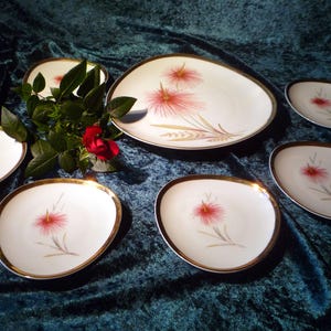 Puede incluir: Un juego de platos de cerámica blanca con bordes dorados, con un diseño floral rosa. Los platos, de varios tamaños y formas, están dispuestos sobre una superficie de terciopelo verde oscuro. Una rosa roja y hojas verdes en el centro.