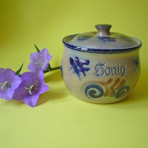 Può includere: Un vasetto di miele in ceramica con coperchio, con disegni floreali blu e la parola "Honig". Il vasetto è beige con accenti blu ed è accompagnato da fiori di campanula viola su sfondo giallo.