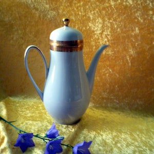 Könnte beinhalten: Eine hellblaue Kaffeekanne aus Porzellan mit hohem, schlankem Körper, geschwungenem Henkel und langem Ausguss. Der Deckel ist mit einem kleinen goldfarbenen Knauf versehen und weist an der Basis ein breites, strukturiertes Goldband auf.