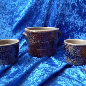 Puede incluir: Tres ollas de cerámica antiguas, en tonos marrones y azules, con texto, sobre una superficie de terciopelo azul. La olla más grande tiene asas y la palabra "Marmelade". Las ollas más pequeñas también tienen texto. Una rosa amarilla en el fondo.