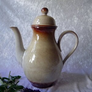 May include: A ceramic coffee pot with a lid, spout, and handle. The pot has a beige body with a brown rim and lid. The handle is curved, and the spout is slender. The pot is likely vintage or handmade.