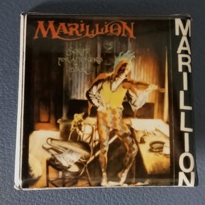 May include: A Marillion album cover featuring a jester in a yellow vest and harlequin pants, holding a violin. The album title "Script for a Jester's Tear" is visible, along with the band's name in red and white lettering.