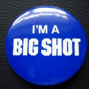 May include: A round, blue button with the text "I'M A BIG SHOT" in bold, white letters. The button has a glossy finish and is set against a textured, black background.