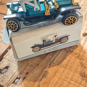 Peut inclure: Une voiture jouet vintage, de couleur teal et noire, avec des détails dorés, posée sur sa boîte assortie. La voiture a un toit blanc et des roues dorées. La boîte porte une image de la voiture et le mot "REO".