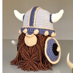May include: A crochet gnome wearing a light gray and blue horned helmet with a beige trim. The gnome has a large beige nose and a long, thick brown yarn beard. It holds a round blue and black shield with a beige border. Text reads crochet pattern PDF.