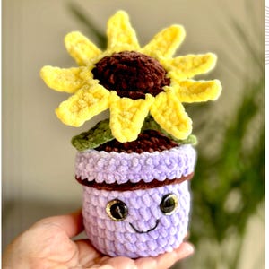 Pode incluir: Um girassol de crochê em um vaso de lavanda com um rosto sorridente e olhos de botão. O girassol tem pétalas amarelas e um centro marrom. O texto "crochet pattern PDF" está à direita.