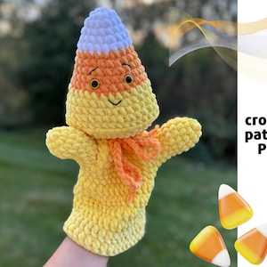 Puede incluir: Títere de maíz de caramelo de felpa hecho a mano con una cara sonriente, un lazo naranja y colores blanco, naranja y amarillo. El títere tiene brazos y está diseñado para usarse en la mano. El texto "crochet pattern PDF" es visible.