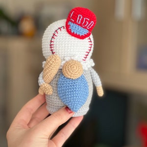 May include: A crocheted gnome wearing a red baseball cap with the word "LOGO" embroidered on it. The gnome is holding a blue crocheted baseball bat.