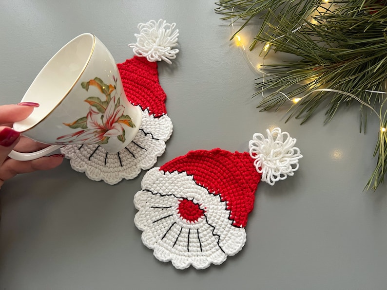 K&ouml;nnte beinhalten: Zwei geh&auml;kelte Weihnachtsmann-Hut-Untersetzer in Rot und Wei&szlig;. Die Untersetzer sind aus Garn gefertigt und haben einen wei&szlig;en Bommel oben drauf.