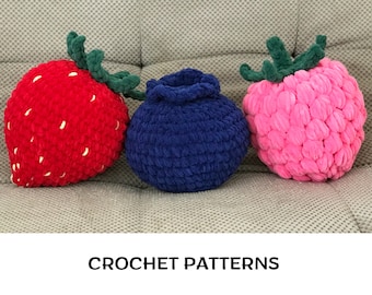 May include: Three crocheted fruit-shaped pillows: a red strawberry, a pink raspberry, and a blue blueberry. The pillows are arranged on a light gray couch. CROCHET PATTERNS is written in black text below the pillows.