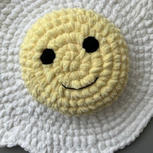 K&ouml;nnte beinhalten: Ein geh&auml;keltes gelbes Kissen mit einem Smiley-Gesicht mit schwarzen Augen und einem L&auml;cheln, das auf einem wei&szlig;en, geh&auml;kelten, eif&ouml;rmigen Kissen liegt.
