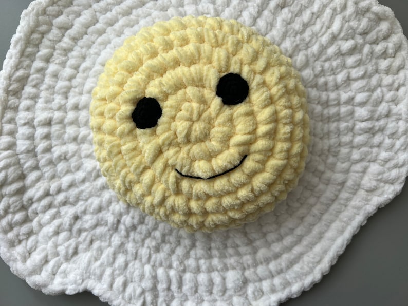 K&ouml;nnte beinhalten: Ein geh&auml;keltes gelbes Kissen mit einem Smiley-Gesicht mit schwarzen Augen und einem L&auml;cheln, das auf einem wei&szlig;en, geh&auml;kelten, eif&ouml;rmigen Kissen liegt.