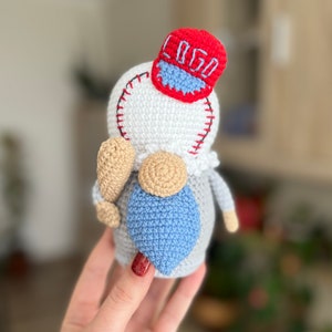 May include: A crocheted gnome wearing a red baseball cap with the word "GO" embroidered on it. The gnome is holding a brown and white baseball bat. The gnome is wearing a white and gray crocheted outfit with a blue crocheted beard.