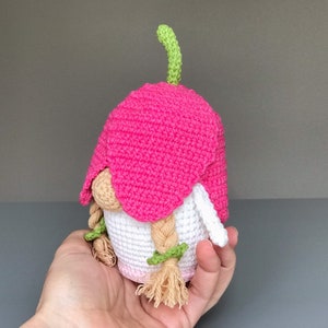 May include: A pink crocheted gnome with a green stem on top of its head. The gnome has white yarn for its body and long blonde yarn for its hair.