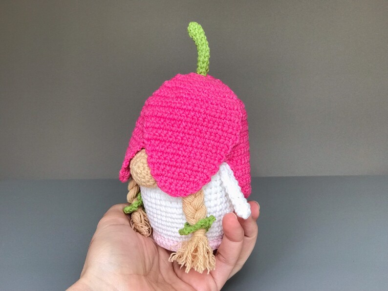 May include: A pink crocheted gnome with a green stem on top of its head. The gnome has white yarn for its body and long blonde yarn for its hair.