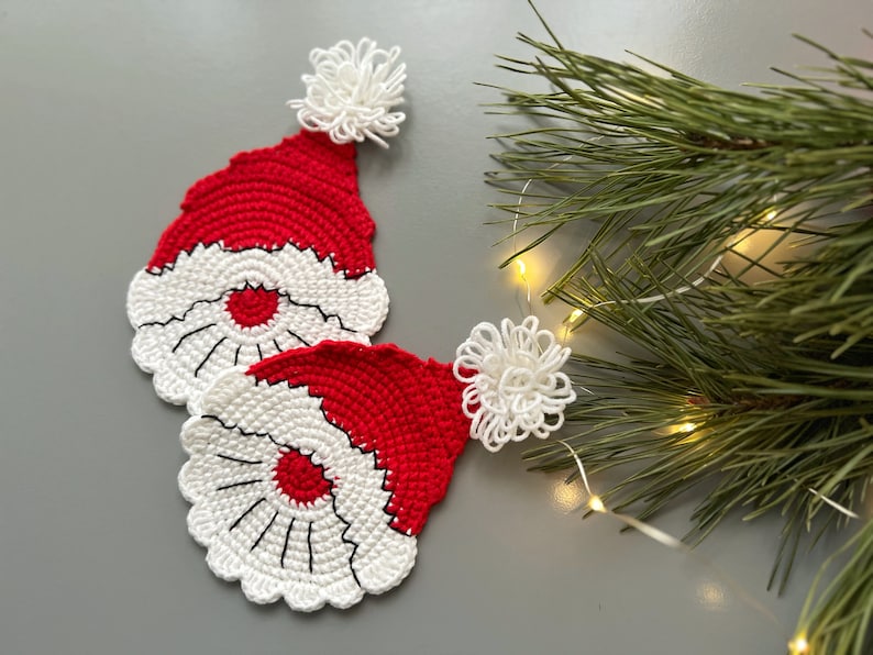 K&ouml;nnte beinhalten: Zwei geh&auml;kelte rote und wei&szlig;e Weihnachtsmann-Hut-Untersetzer mit wei&szlig;en Bommeln. Die Untersetzer befinden sich auf einer grauen Oberfl&auml;che mit einem Kiefernzweig und Lichtern im Hintergrund.