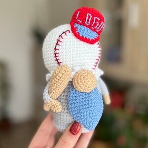 May include: A crocheted gnome wearing a red baseball cap with the word "LOGO" embroidered on it. The gnome is holding a wooden baseball bat and has a blue crocheted beard.