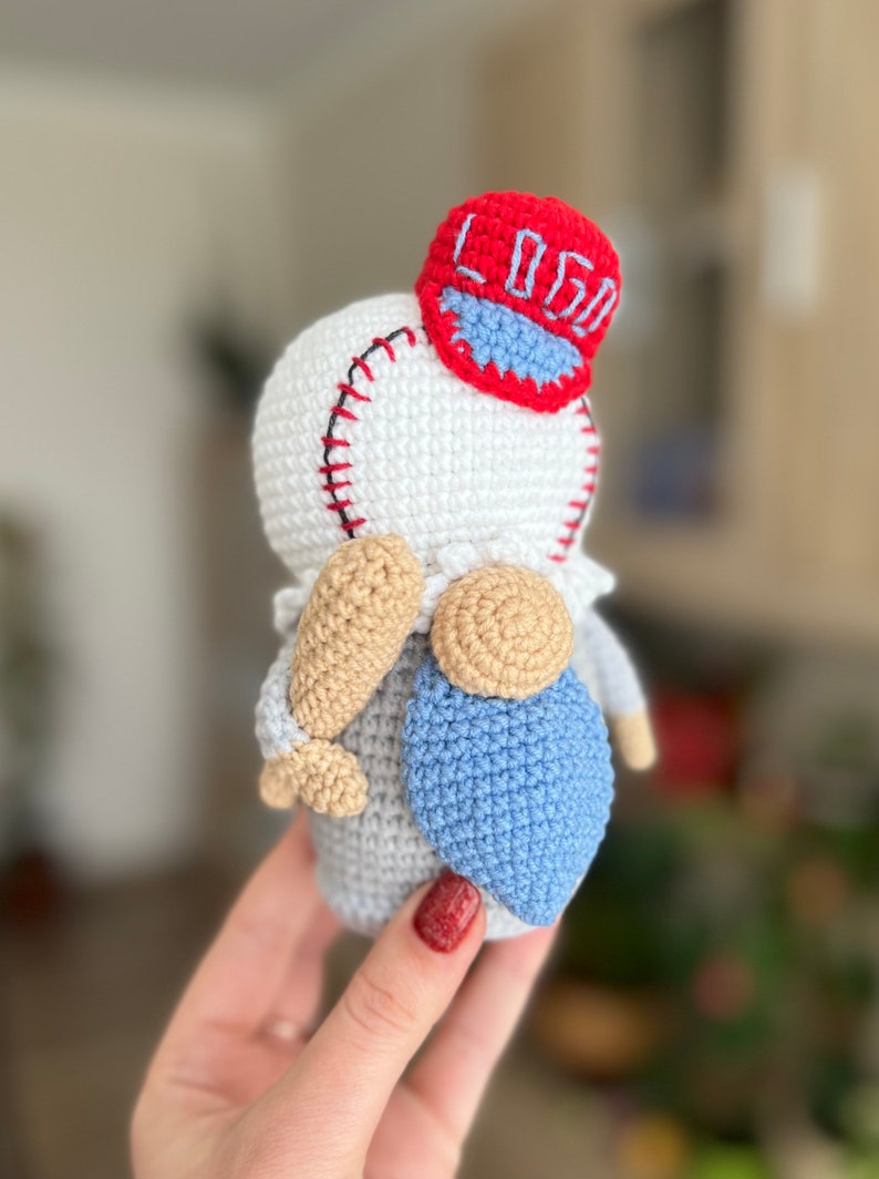 May include: A crocheted gnome wearing a red baseball cap with the word "LOGO" embroidered on it. The gnome is holding a wooden baseball bat and has a blue crocheted beard.