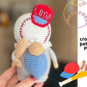 May include: Crochet pattern PDF for a baseball-themed gnome wearing a red and white hat with the word "LOGO" on it. The gnome is holding a brown baseball bat and has a blue heart on its chest.
