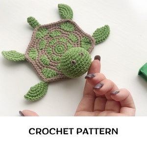 May include: A crocheted green and brown turtle with a shell that has a circular pattern. The turtle is lying on a white surface.
