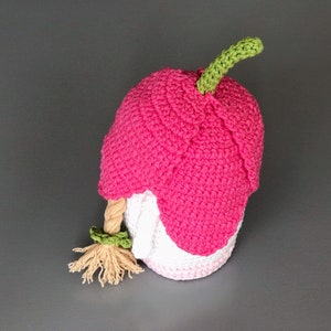 May include: A pink crocheted gnome with a green stem on top of its head and a long blonde braid. The gnome is wearing a white crocheted hat.
