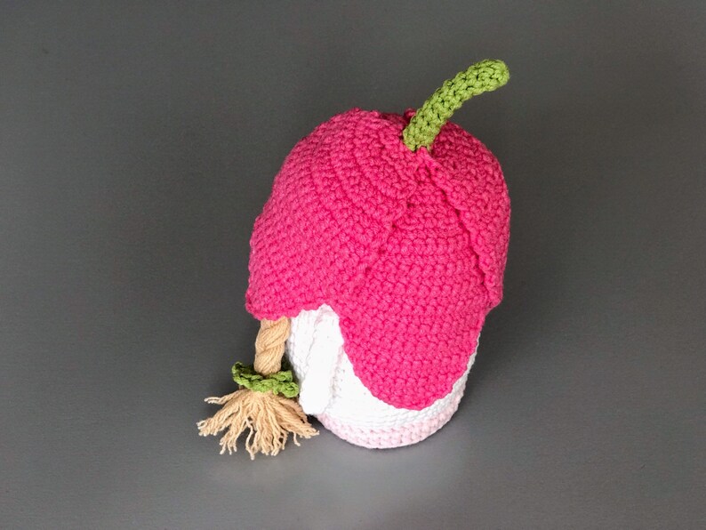 May include: A pink crocheted gnome with a green stem on top of its head and a long blonde braid. The gnome is wearing a white crocheted hat.