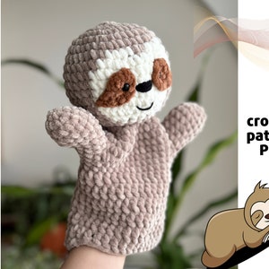Puede incluir: Un títere de perezoso de crochet con pelaje marrón, ojos blancos y nariz marrón. El títere está siendo sostenido por una mano, y el fondo es una imagen borrosa de una planta.