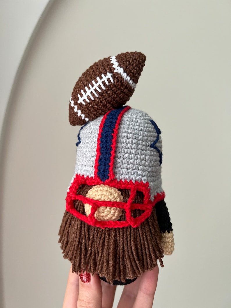 May include: Crocheted gnome wearing a football helmet and holding a football. The gnome has a brown beard and a red nose.