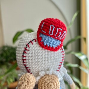 May include: A crocheted doll wearing a red and blue baseball cap with the word "Loso" embroidered on it. The doll has a white body, tan arms and legs, and a blue and white striped shirt.