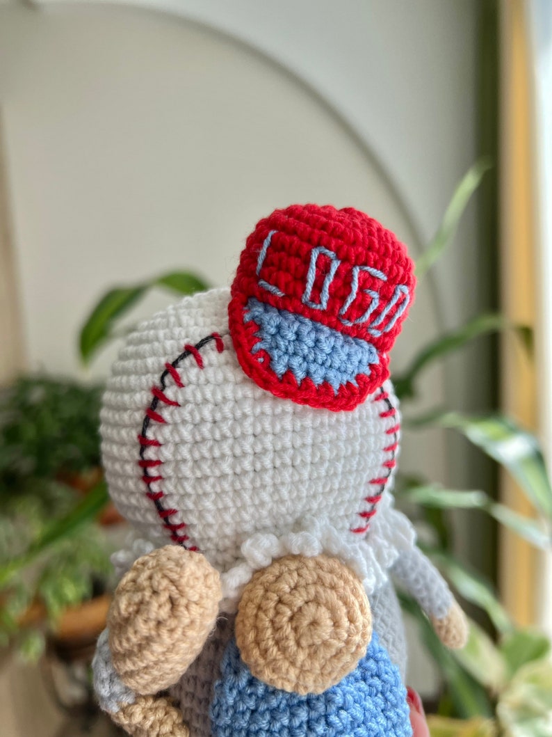 May include: A crocheted doll wearing a red and blue baseball cap with the word "Loso" embroidered on it. The doll has a white body, tan arms and legs, and a blue and white striped shirt.
