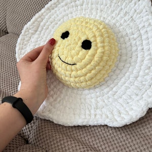 K&ouml;nnte beinhalten: Ein wei&szlig;es, geh&auml;keltes Kissen in Eierform mit einem gelben, geh&auml;kelten Smiley-Gesicht in der Mitte.