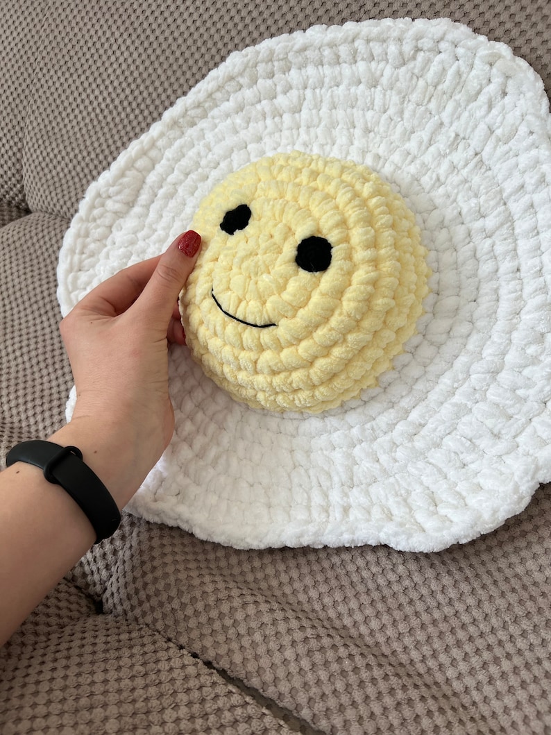 K&ouml;nnte beinhalten: Ein wei&szlig;es, geh&auml;keltes Kissen in Eierform mit einem gelben, geh&auml;kelten Smiley-Gesicht in der Mitte.