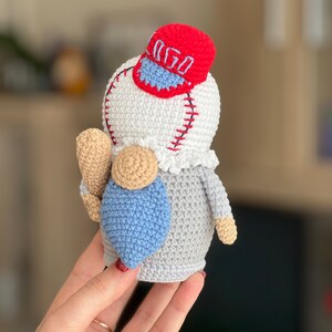 May include: A crocheted gnome wearing a red baseball cap with the word "GO" embroidered on it. The gnome is holding a baseball bat and has a blue and gray body.