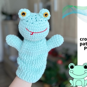 May include: A light blue crocheted frog puppet with yellow eyes and a red tongue. The puppet is being held by a hand. The image also includes the text "crochet pattern PDF".