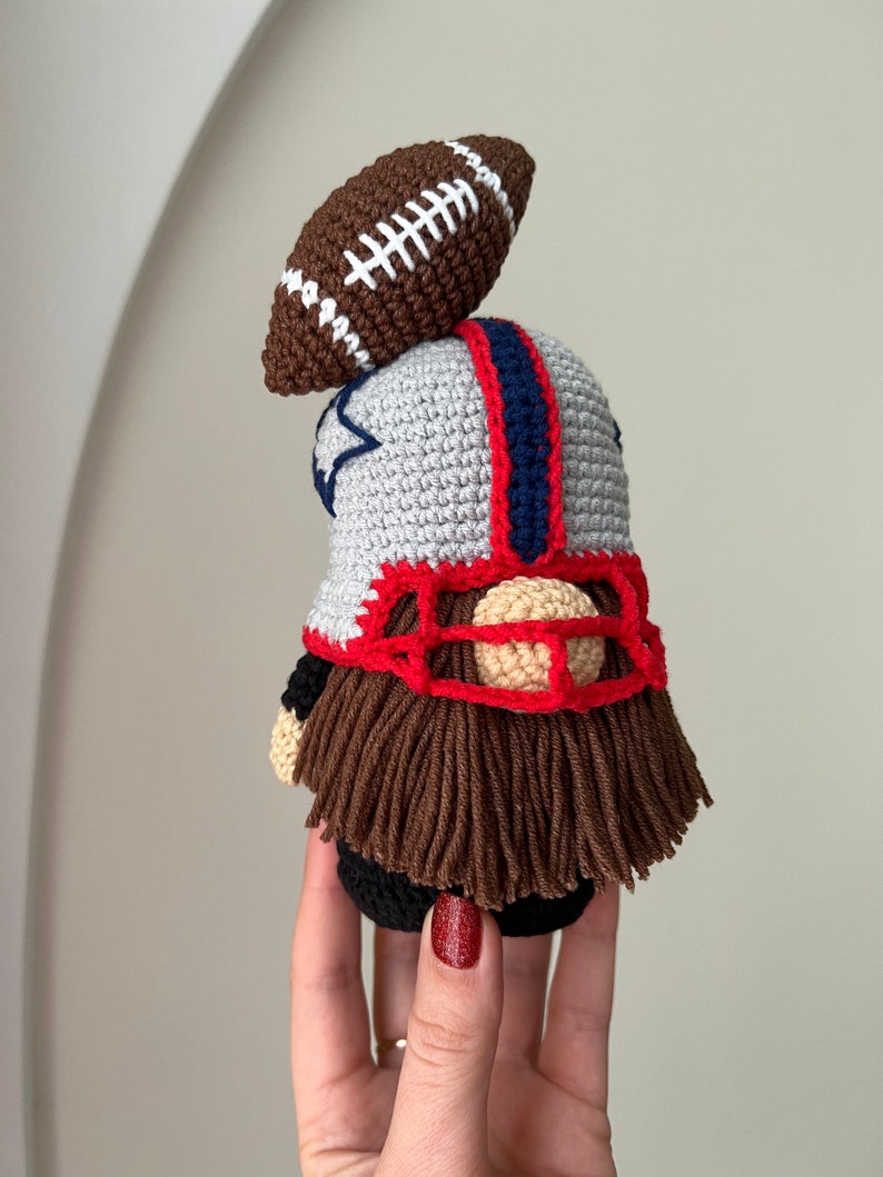 May include: A crocheted gnome wearing a football helmet and holding a football. The gnome has a brown beard and is wearing a gray, red, and blue uniform.
