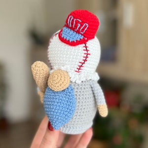May include: A crocheted baseball gnome wearing a red baseball cap with the word "GO" on it. The gnome is holding a brown baseball bat and has a blue and gray crocheted body.