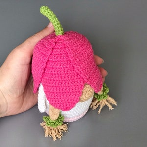 May include: A pink crocheted gnome with a green stem on top of its hat. The gnome has a white crocheted body and brown yarn hair.