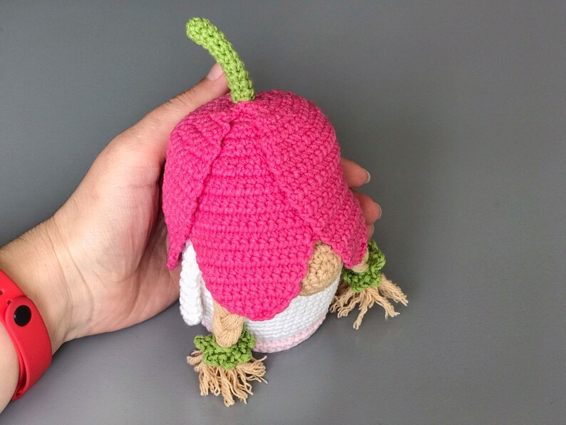May include: A pink crocheted gnome with a green stem on top of its hat. The gnome has a white crocheted body and brown yarn hair.