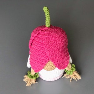 May include: A pink crocheted gnome with a green stem on top of its head. The gnome has a white crocheted body and brown yarn hair.