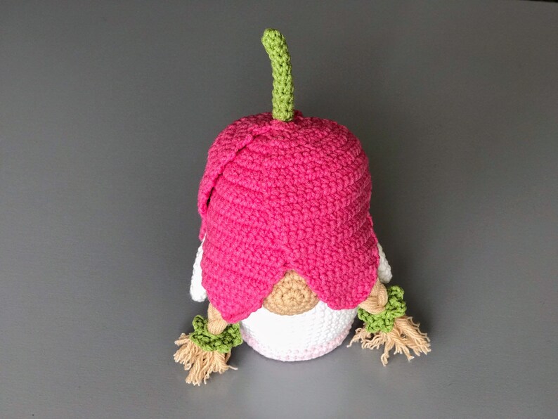 May include: A pink crocheted gnome with a green stem on top of its head. The gnome has a white crocheted body and brown yarn hair.
