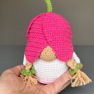 May include: A crocheted gnome doll with a pink flower hat and green stem, white body, and blonde braided hair with green bows.