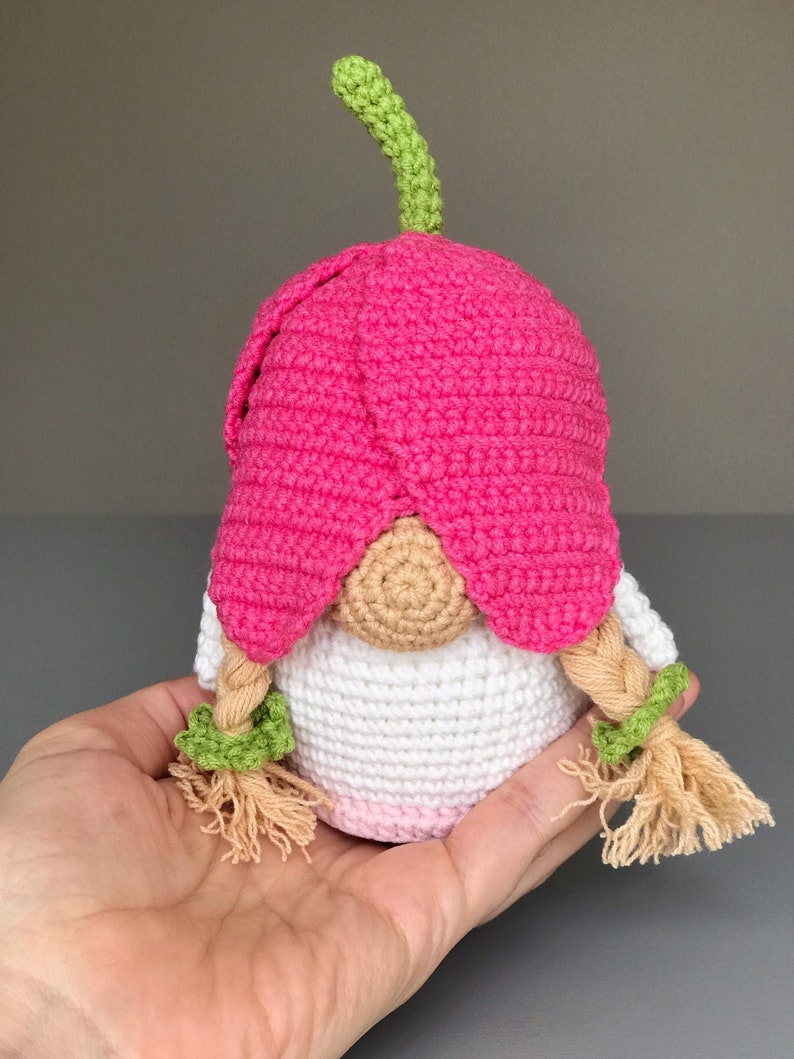 May include: A crocheted gnome doll with a pink flower hat and green stem, white body, and blonde braided hair with green bows.